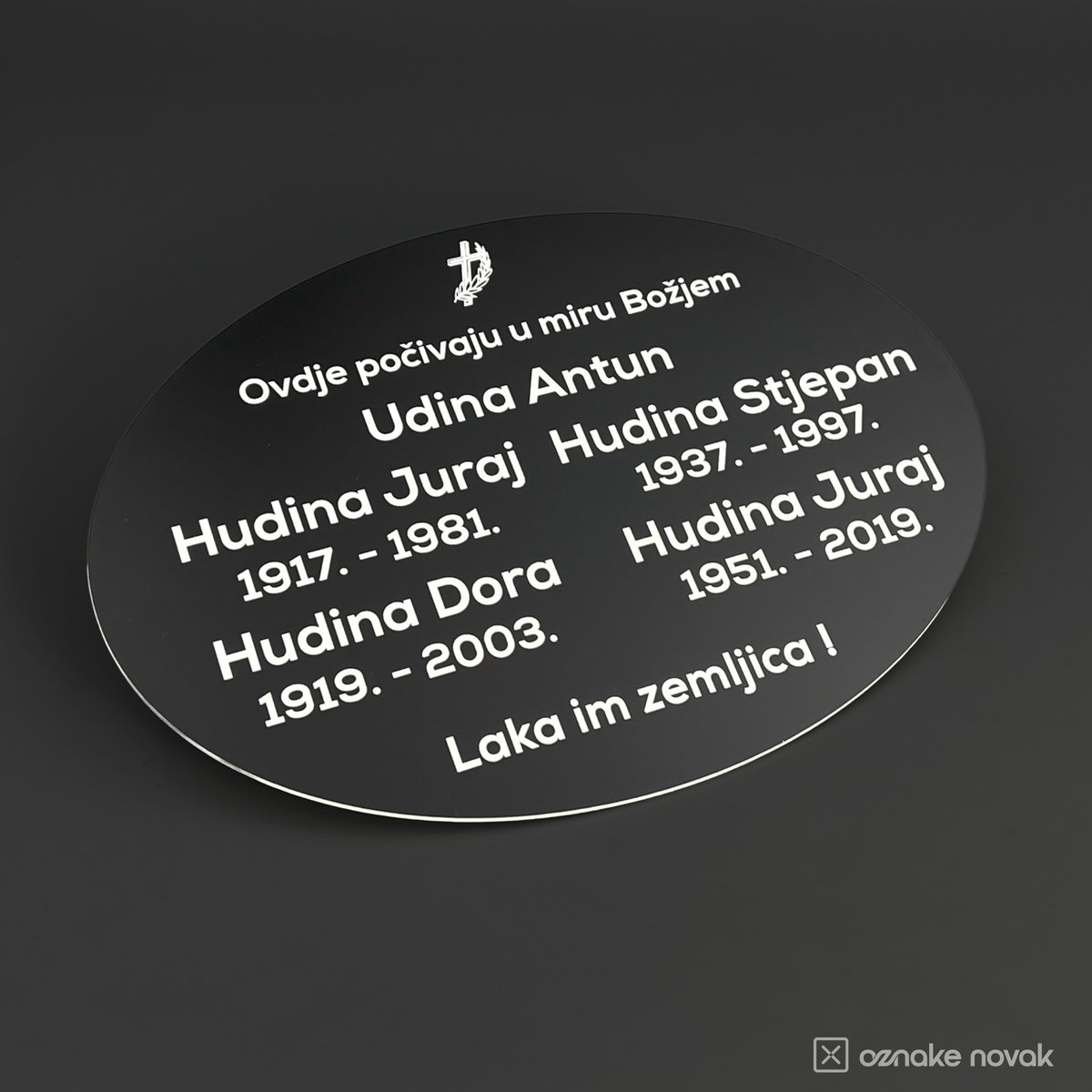 Black oval memorial plate with white engraved names, dates and a cross, angled view