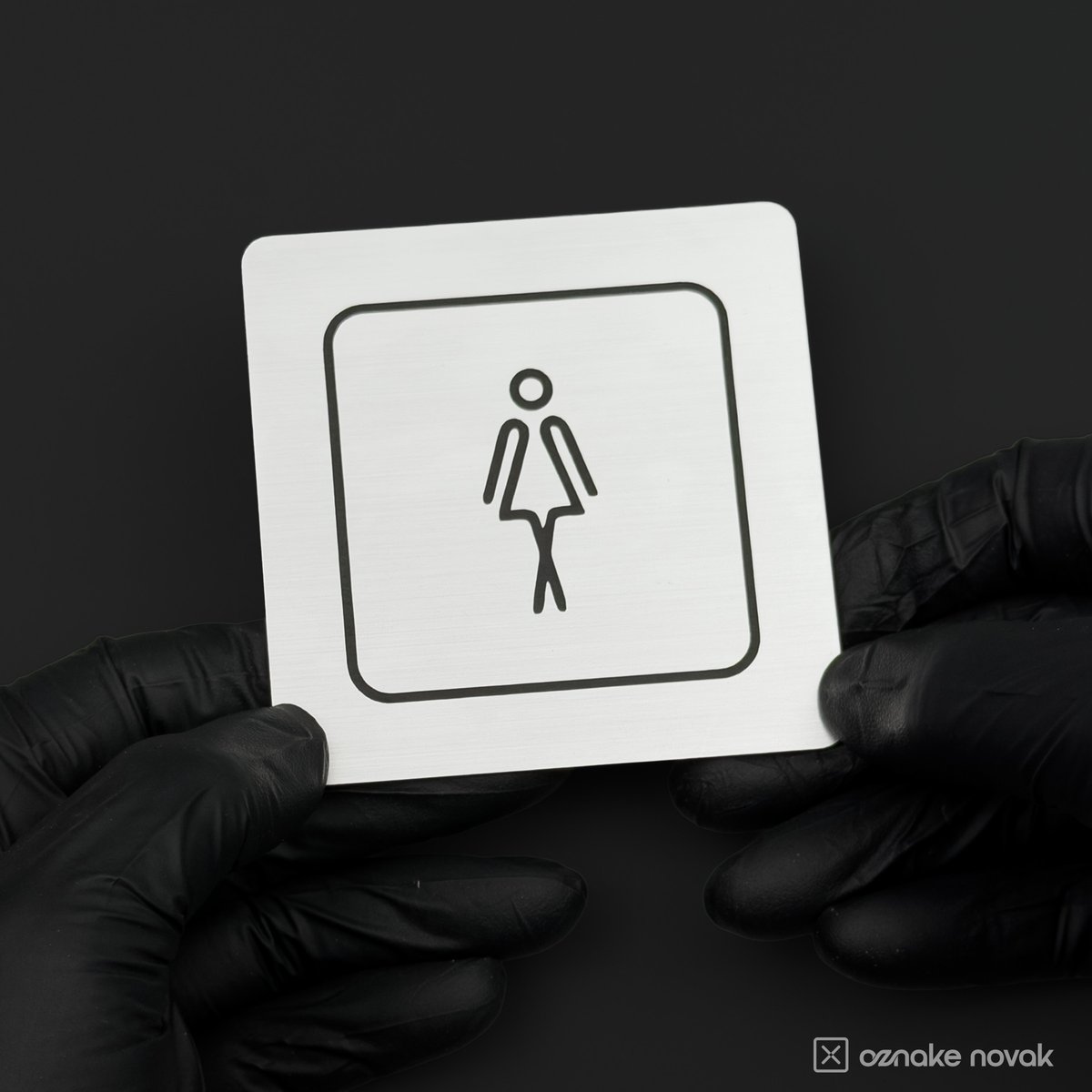 Gloved hands holding a brushed silver restroom sign with women's pictogram