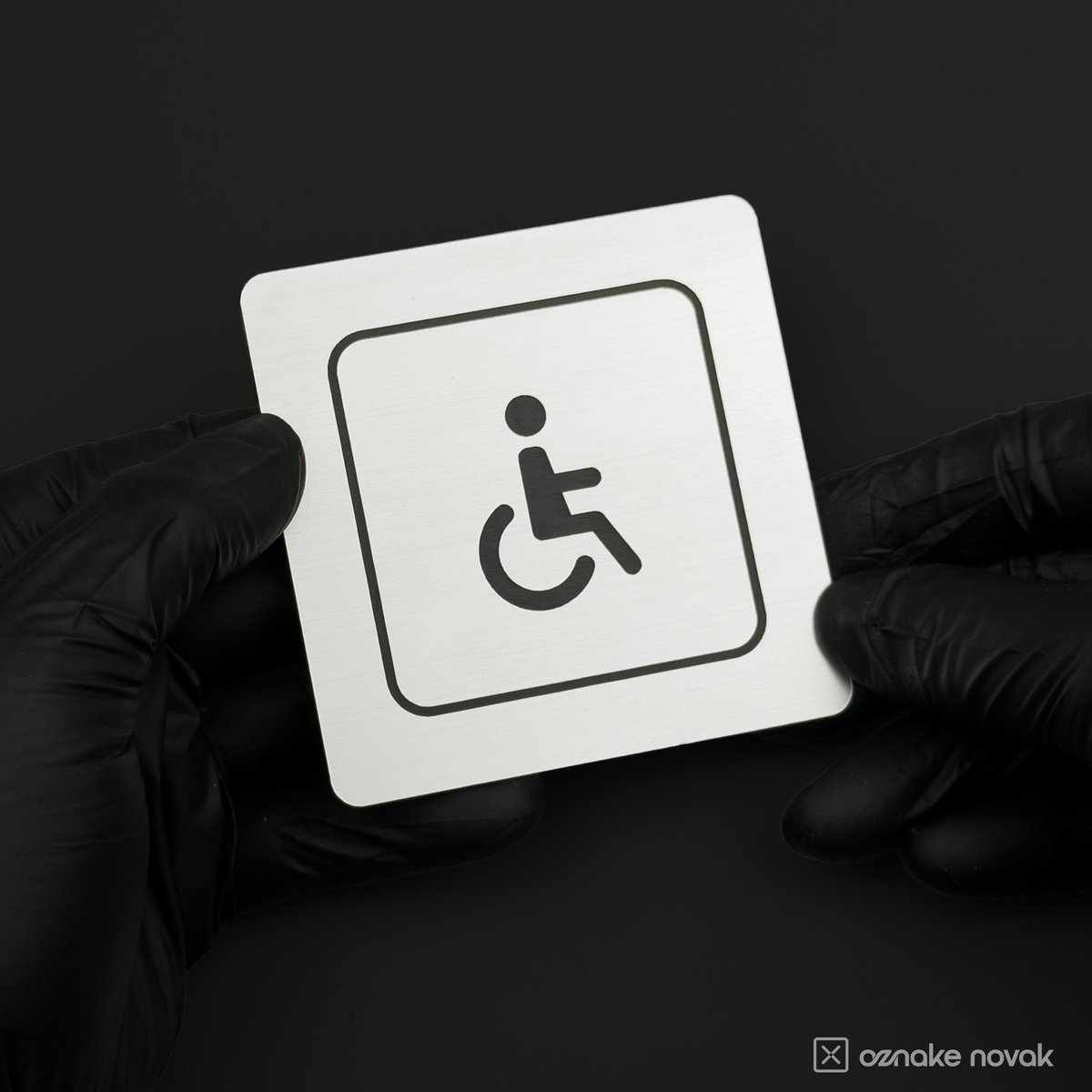 Gloved hands holding a brushed silver restroom sign with accessible pictogram