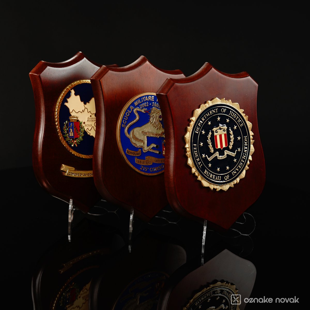 Three wooden shield plaques with enamelled emblems standing side by side on clear stands